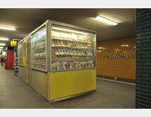 Ein Kiosk in einer U-Bahn-Station, umgeben von einem Gittergehäuse mit gelbem unteren Bereich, das Zeitschriften und Zeitungen ausstellt. Im Hintergrund sind orange geflieste Wände, eine Leuchtreklame und ein Schild mit der Aufschrift „Gontzelstrasse“ zu sehen.