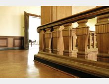 Innenansicht eines historischen Gebäudes mit einer Holzbrüstung aus kunstvoll verzierten Balustern. Im Hintergrund ist eine geöffnete Tür zu einem hell erleuchteten Raum mit Stühlen zu sehen.