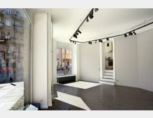 Ein leerer, heller Innenraum mit großen Schaufenstern, die den Blick auf eine städtische Straße mit mehrstöckigen Gebäuden ermöglichen. Der Raum hat weiße Wände, einen grauen Boden und eine schwarze Schienenbeleuchtung an der Decke, mit einer kleinen Treppe, die in einen angrenzenden Küchenbereich führt.