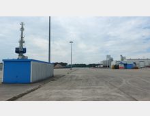 Industriegelände mit großen, leeren Betonflächen, einer blauen Maschinenhalle links und einem hellen Industriegebäude rechts. Im Hintergrund ist ein Stapel Rohmaterial zu erkennen, umgeben von Bäumen und Freiflächen.