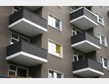 Mehrfamilienhaus mit grauer Fassade und hervorstehenden Balkonen, die schwarze Kacheln und weiße Kanten haben, neben rechteckigen Fenstern.