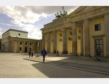 Das Bild zeigt das Brandenburger Tor in Berlin, ein neoklassizistisches Denkmal mit großen Säulen und der Quadriga-Statue auf dem Dach. Im Vordergrund befindet sich ein Platz mit Menschen, die zu Fuß oder mit Fahrrädern unterwegs sind, bei leicht bewölktem Himmel.