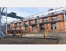 Backstein-Gebäude einer Industrieanlage mit großen Fenstern und einem angrenzenden Gelände, das mit Metallträgern, Rohren und einer Krananlage ausgestattet ist. Im Hintergrund ragt ein roter Wasserturm empor.