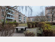 Mehrstöckige Wohngebäude mit verputzten Fassaden und Balkons umgeben einen Innenhof mit Gartenanlage. Im Vordergrund sind eine Tischtennisplatte, eine Bank und kahle Büsche sichtbar, die den gepflasterten Bereich säumen.