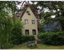 Einfamilienhaus im Landhausstil mit hellem Putz, roten Fensterrahmen und einem steilen Ziegeldach, umgeben von Bäumen und Sträuchern; im Vordergrund ein kleiner Garten mit einer Bank.