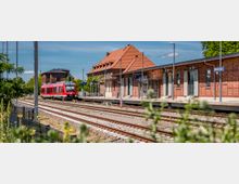Bahnhof mit mehreren Gleisen und einem Bahnsteig vor einem langen Backsteingebäude mit rotem Ziegeldach, Lampenmasten und Beschilderung. Links steht ein roter Regionalzug an den Gleisen, umgeben von Grünflächen und Bäumen.