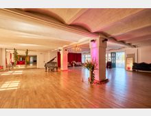 Großer, offener Veranstaltungsraum mit Holzparkett, niedriger Decke und weißen Stützpfeilern mit farbiger Beleuchtung. Im Raum stehen ein Flügel, ein Kronleuchter, rote Vorhänge sowie Sitzmöbel; große Fenster und eine geöffnete Tür lassen Tageslicht herein.