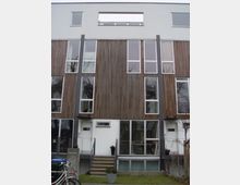 Moderne Reihenhausfassade mit Holzlamellen und großen Glasfronten. Die oberen Stockwerke bestehen aus einer Kombination aus weißen Putzflächen, bodentiefen Fenstern und einem schlichten Design, während der Eingangsbereich kleine Treppen und vereinzelt Pflanzen in Töpfen aufweist.