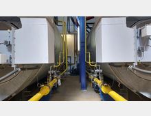 Zwei große industrielle Kessel in einem technischen Raum, verbunden durch gelbe Gasleitungen mit Ventilen und Rohrsystemen. Die Umgebung ist sauber mit Betonboden, blauer Metallstütze und Wänden aus weißem Mauerwerk.