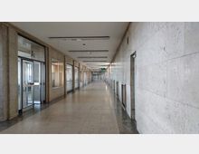 Langer, heller Korridor in einem Bürogebäude mit Wänden aus hellem Stein und Bodenfliesen. Links sind Glastüren und Fensterrahmen zu sehen, rechts eine durchgehende Steinwand mit kleinen Wandöffnungen.