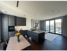 Offene, moderne Wohnung mit dunkler Einbauküche, hellem Esstisch und integrierter Wohnzimmerecke. Große Glas-Schiebetüren führen zu einer Terrasse mit Blick auf die Stadt.