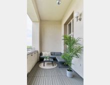 Überdachter Balkon mit grauem Bodenbelag, einer Sitzgruppe aus grauem Sofa und Tisch, dekoriert mit Kissen und kleinen Pflanzen. Eine Topfpflanze steht rechts, und an der Decke hängt eine glänzende Kugel.