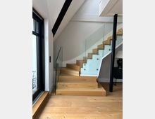 Moderner Innenbereich mit einer Holztreppe, die nach rechts oben führt, und einem seitlichen Glasgeländer mit Metallhaltern. Links befindet sich eine große Glastür oder Fensterfront, darunter ein Holzfußboden; rechts ist ein schwarzer Stützpfosten zu sehen.