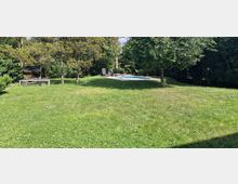 Weitläufiger Garten mit Rasenfläche, mehreren Bäumen und Sträuchern. Im Hintergrund befindet sich ein kleiner Pool mit Liegestühlen und einem Sonnenschirm, umgeben von dichtem Grün.