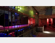 Ein industriell gestalteter Innenraum mit Betonwänden und Decke, durchzogen von metallenen Gitterstäben, die einen Bereich abtrennen. Bunte Lichteffekte (Rot, Blau, Grün) erleuchten den Raum, und im Hintergrund sind eine Backsteinmauer und eine Tür sichtbar.
