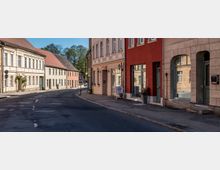 Curving paved street in a small town lined with two-story historic buildings in light stucco and brick, featuring arched doorways and shopfront windows. A sidewalk with street lamps and a “30 zone” traffic sign runs along the buildings, with trees visible at the far end of the road.