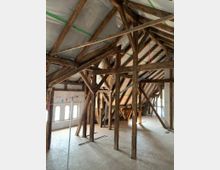 Ein Dachboden mit sichtbaren Holzbalken und einer teilweise renovierten Struktur. Der Raum hat mehrere Fenster mit Rundbögen auf einer Seite und Holzbodenplatten.