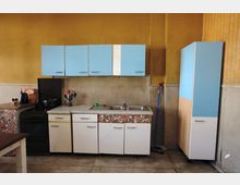 Kleine Küche mit gelben Wänden, blauen Oberschränken und weißen Unterschränken mit bunten Mustern. Die Küche enthält ein Spülbecken, einen Herd, eine Kaffeemaschine und Reinigungsutensilien; der Boden besteht aus hellen Fliesen.