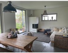 Wohnzimmer mit modernem Interieur: Im Vordergrund ein Holz-Esstisch mit Kerzen, im Hintergrund eine helle Eckcouch, ein Kamin und ein großes Fenster mit Gartenblick. Der Raum ist minimalistisch gestaltet und hat Betonboden sowie dezente Dekorationen.