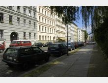 Straßenansicht mit Wohnhäusern im Altbaustil, die mit hellen Fassaden und hohen Fenstern gestaltet sind. Am Straßenrand stehen geparkte Autos, daneben ein gepflasterter Gehweg mit vereinzelten Bäumen und einer Laterne.
