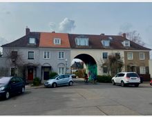 Wohnstraße mit einem mehrstöckigen Reihenhaus, dessen mittlerer Teil einen großen, bogenförmigen Durchgang mit bemalter Wand zeigt. Vor dem Gebäude parken Autos, und zwei Radfahrer fahren durch die Straße.