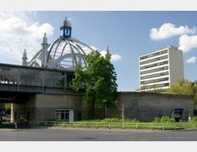 Eine erhöhte U-Bahn-Station mit einer markanten gläsernen Kuppel, gekrönt von einem großen blauen „U“-Symbol. Im Vordergrund sind Bäume, ein Fußgängerweg, sowie ein Teil der Straße mit einem Verkehrsschild sichtbar, im Hintergrund ein mehrstöckiges Bürogebäude.