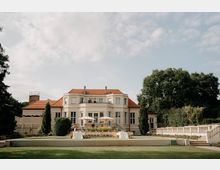 Große, helle Villa mit rotem Ziegeldach und symmetrischer Fassade, davor eine breite Terrasse mit gedeckten Tischen und Sonnenschirmen. Im Vordergrund liegt eine weitläufige Rasenfläche, seitlich eingefasst von Bäumen, Hecken und einer Balustrade.