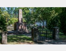 Denkmal auf einem parkähnlichen Gelände mit einem Obelisken aus Stein, verziert mit einer goldenen Sternsymbolik und einer Inschrift. Im Vordergrund eine niedrige metallene Eingangspforte mit roten Sternen, umgeben von Bäumen und Gebäuden im Hintergrund.