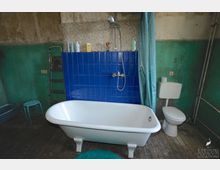 Ein einfaches Badezimmer mit einer freistehenden weißen Badewanne auf einem Holzboden, einer blauen gefliesten Duschwand mit Duscharmatur und einem türkisfarbenen Duschvorhang. Im Hintergrund befinden sich eine Toilette, eine grün gestrichene Wand sowie diverse Alltagsgegenstände auf der Wandablage.