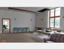 Ein leerer Raum mit abgenutztem Teppichboden und sichtbaren Schäden an der Decke. Im Raum stehen blaue Stühle an der Wand, ein Tisch mit Büchern und Kartons sowie ein altes Sofa. Große Fenster lassen Tageslicht herein, während im Hintergrund ein Durchgang in einen angrenzenden Raum zu sehen ist.