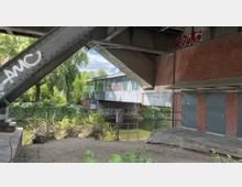 Ein Blick unter eine Brücke, die aus Stahlträgern und Backstein gebaut ist, mit Graffiti an den Wänden. Darunter verläuft ein Fluss, umgeben von dichter Vegetation, und im Hintergrund ist ein Gebäude mit Glasfassade und weiteren Graffiti zu sehen.