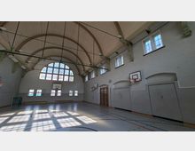 Innenansicht einer großen Turnhalle mit hoher Tonnengewölbe-Decke, Oberlichtfenstern und einem großen Rundbogenfenster an der Stirnseite. An den hellgrauen Wänden hängen mehrere Basketballkörbe; auf dem Hallenboden sind Spielfeldlinien markiert und seitlich stehen Bänke sowie eine Sprossenwand.