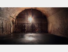 Gewölbekeller mit rustikalen Ziegelwänden und einer gewölbten Decke aus Backstein. Ein einzelnes hängendes Licht beleuchtet den dunklen Raum, der einen feuchten Boden und befestigte Metallrohre an den Wänden aufweist.