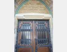 Holz-Doppeltür mit verzierten Eisengittern in einem historischen Gebäude, darüber ein steinernes Rundbogenportal mit goldener Inschrift auf grünem Hintergrund. Die Architektur weist neugotische Elemente auf.