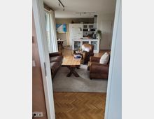 Wohnraum mit offener Küche, ausgestattet mit zwei braunen Ledersesseln, einem Holztisch auf einem grauen Teppich sowie einer weißen Küchenzeile im Hintergrund. Große Fenster lassen Tageslicht herein, und es sind minimalistische Dekoelemente sichtbar.