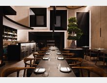 Modernes Restaurant mit minimalistischer Einrichtung, einer langen, gedeckten Tafel mit Holzstühlen und zentral platziertem Bonsai-Baum in einem erhöhten Pflanzgefäß. Im Hintergrund befinden sich eine dunkel gehaltene Bar mit beleuchteten Regalen und geometrischen Wandvertiefungen.