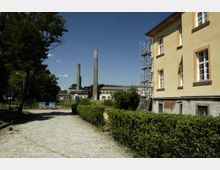 Gepflasterter Weg mit Hecken, der an einem gelben Gebäude mit Gerüst vorbeiführt; im Hintergrund sind hohe Industrieschornsteine und ein Fabrikgebäude zu sehen, eingerahmt von Bäumen unter klarem blauem Himmel.