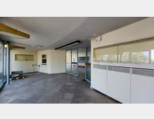 Leerer Eingangsbereich eines Gebäudes mit gefliesten Böden und bräunlich-beigen Wänden. Große Glastüren mit orangenen Griffen führen in den Innenbereich, links befinden sich Paletten und Baustellenelemente, oben sind Deckenleuchten und teilweise abblätternder Putz sichtbar.