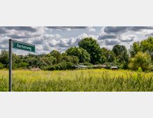 Grüne Wiesen mit hohen Pflanzen im Vordergrund, dahinter mehrere kleine Gartenhäuser, umgeben von dichtem Baumbestand bei bewölktem Himmel. Links im Bild ein Straßenschild mit der Aufschrift „Gartenweg“.