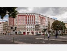 Eckgebäude mit historischer Fassade in Weiß und Rot an einer Straßenkreuzung in einem städtischen Bereich, im Erdgeschoss befinden sich Geschäfte, darunter eine Apotheke. Im Vordergrund sind Fußgängerüberwege, Ampeln und parkende Autos zu sehen, flankiert von Gehwegen und Bäumen.