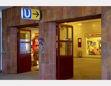 Eingang zu einer U-Bahn-Station mit gelber Wandverkleidung und einem blauen „U“-Schild mit Richtungspfeil darüber. Links im Bild ist ein roter Türrahmen, durch den ein Geschäft mit Kleidungsstücken sichtbar ist, während rechts ein Friseursalon mit beleuchtetem Innenraum zu sehen ist.