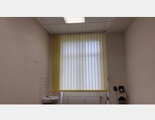 Ein schlichtes Zimmer mit cremefarbenen Wänden, einem großen Fenster mit vertikalen gelben Jalousien und einer weißen Deckenleuchte. An der linken Wand befindet sich ein kleines Waschbecken mit weißen Fliesen, darunter ein Heizkörper; rechts sind technische Geräte auf einem Regal sichtbar.