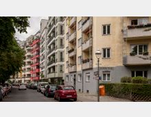 Straßenszene mit mehrstöckigen Wohngebäuden, teils mit Balkonen und gefärbten Fassaden in Beige, Grün, und Rot. Am Straßenrand parken Autos, und im Vordergrund steht eine Straßenlaterne sowie ein Straßenschild mit der Aufschrift „Krefelder Straße“.