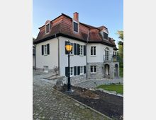 Zweistöckiges Wohnhaus mit weißer Fassade, grauen Fensterläden und rotem Mansardendach. Vor dem Gebäude führt ein gepflasterter Weg vorbei, und eine schwarz lackierte Laterne ist im Vordergrund sichtbar.