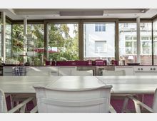 Moderne Küche mit großen bodentiefen Fenstern, die den Blick auf einen begrünten Außenbereich mit Bäumen und einem weißen Gebäude erlauben. Der Raum verfügt über weiß-pinke Küchenschränke, Arbeitsflächen in Magenta und einen großen weißen Esstisch mit passenden Kunststoffstühlen im Vordergrund.