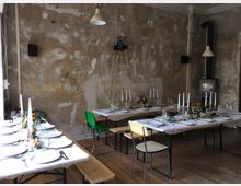 Ein rustikal eingerichteter Raum mit Betonwänden und Holzboden, in dem mehrere Tafeln mit weißen Tischdecken, Kerzenständern und Blumen dekoriert sind. Die Bestuhlung besteht aus verschiedenen Stühlen und Bänken, und an der Wand ist ein Ofen sichtbar.