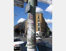 Straßenszene mit einer beklebten Laternenstange im Vordergrund, auf der Zettel und Aufkleber angebracht sind. Im Hintergrund sind mehrstöckige Wohn- und Geschäftsgebäude, parkende Autos und eine Kreuzung bei sonnigem Wetter zu sehen.