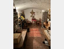 Kleiner Kellerraum mit gewölbter Decke, Backsteinboden und rustikaler Einrichtung. Im Mittelpunkt steht ein kleiner Tisch mit einer roten Tischdecke, umgeben von Stühlen, flankiert von Weinregalen und Holzmöbeln.