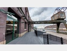 Ein historisches Lagerhaus aus Backstein mit großen Fenstern und grünem Eingang, gelegen an einem Kanal. Rechts im Bild ist ein alter Industriekran aus Metall sowie Lagergebäude und weiße Tanks sichtbar.