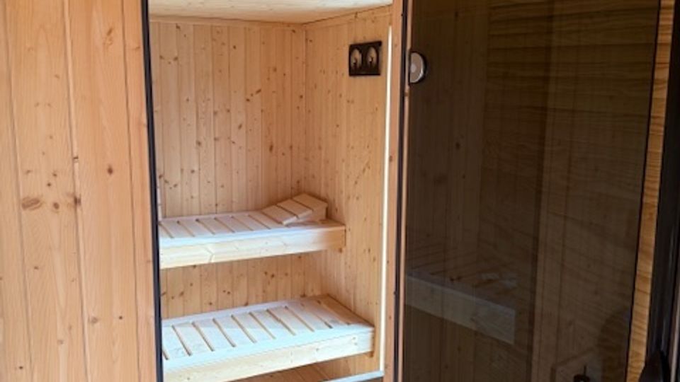 Wood-paneled sauna room with a wooden plank floor and ceiling, viewed through a glass door with a vertical handle. Inside are two-tier wooden bench seats along the back wall and a small heater unit near the entrance.